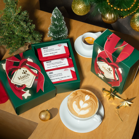 Top view of open and closed green Holiday Trio Coffee boxes, with small pine trees, star ornaments, a latte in a white cup on saucer and an espresso in a white cup on saucer all placed on table.