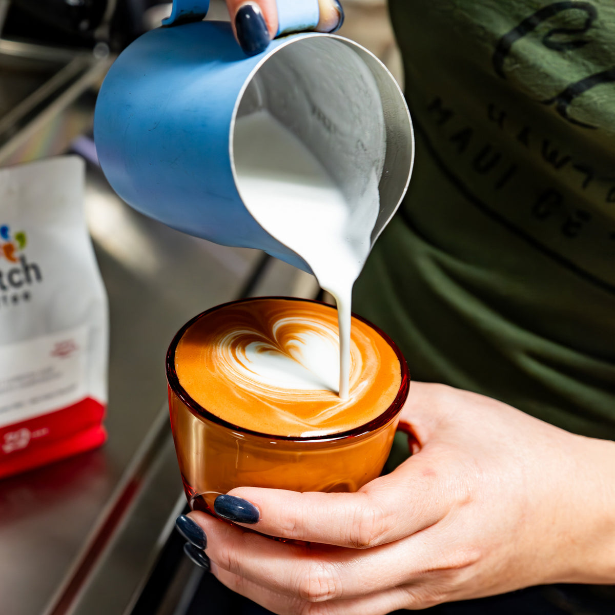 Person pouring milk into a coffee cup with latte art, with a bag of coffee in the background.