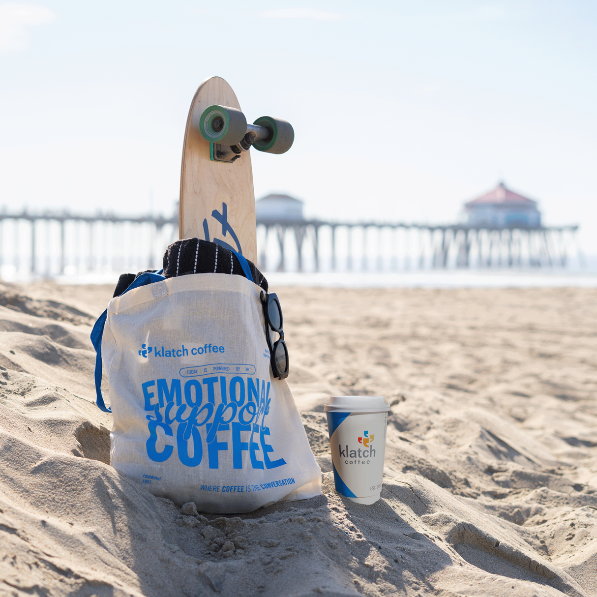 Skateboard with a bag and cup on a sandy beach with a Huntington Pier in the background