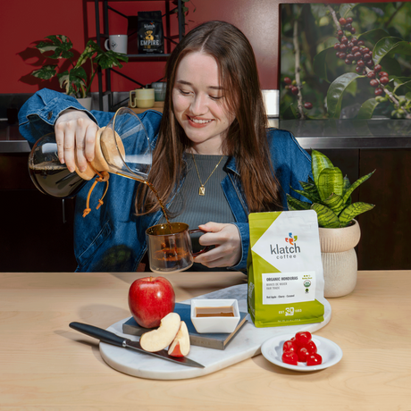 A woman in a kitchen setting pours coffee from a glass pour-over maker into a glass cup, with a bag of Klatch Coffee and coffee beans on the table in front of her.