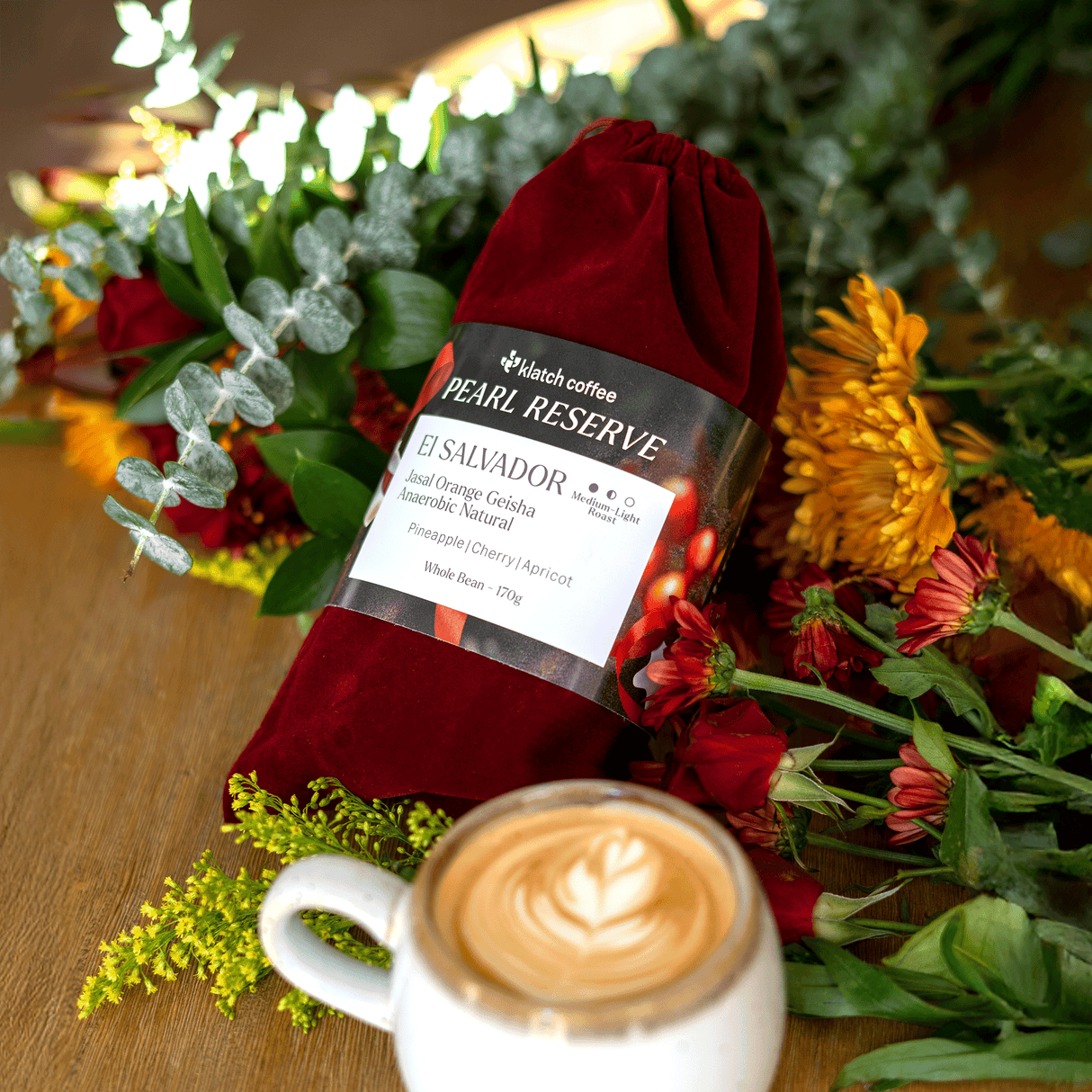 One by one photo of El Salvador Orange Geisha in a red velvet bag on a table, surrounded by flowers and a latte.