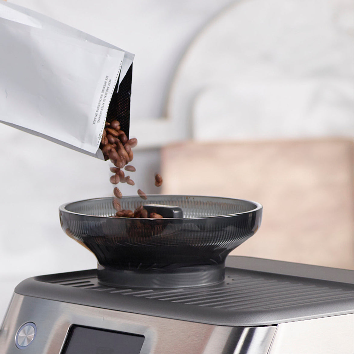 Person pouring coffee beans from a bag into a black hoper of the coffee grinder on the Breville Barista Impress Touch Espresso Machine.