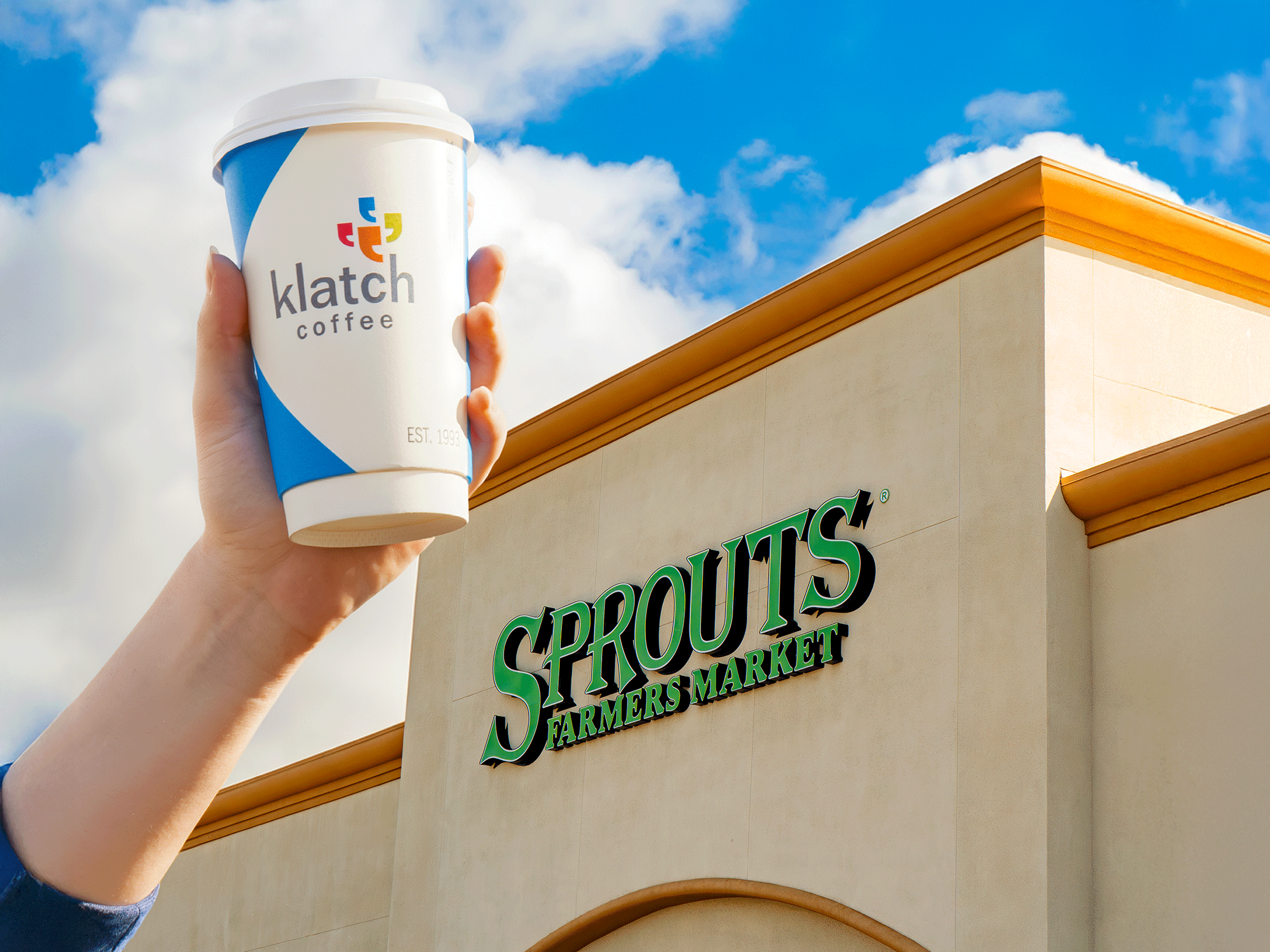 Hand holding a blue Klatch Coffee cup in front of a Sprouts Farmers Market store sign.