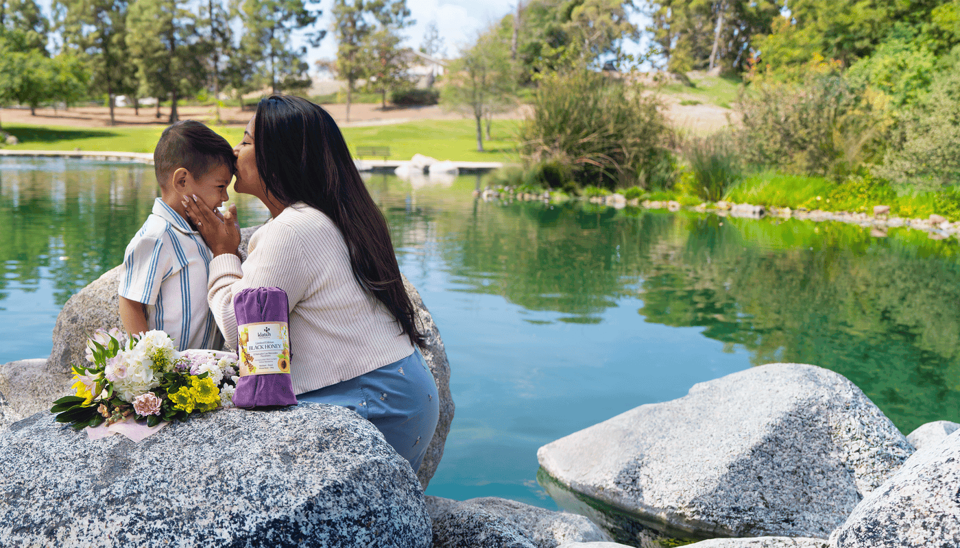 Mother kissing her son’s head at a lake, with Klatch Coffee’s Black Honey Pearl Reserve Coffee in the foreground beside flowers.