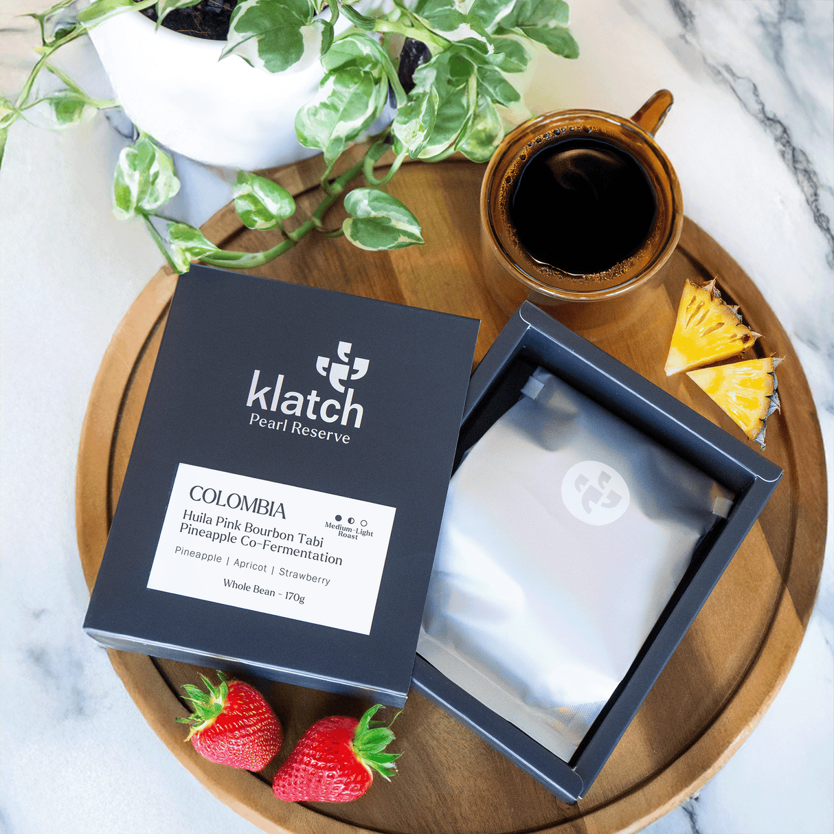 Colombia Huila Pink Bourbon Tabi Pineapple Co-Fermentation on wooden try with glass amber mug of coffee, wedges of pine apple and strawberries place on a white marble counter with small white potted plant top-view.