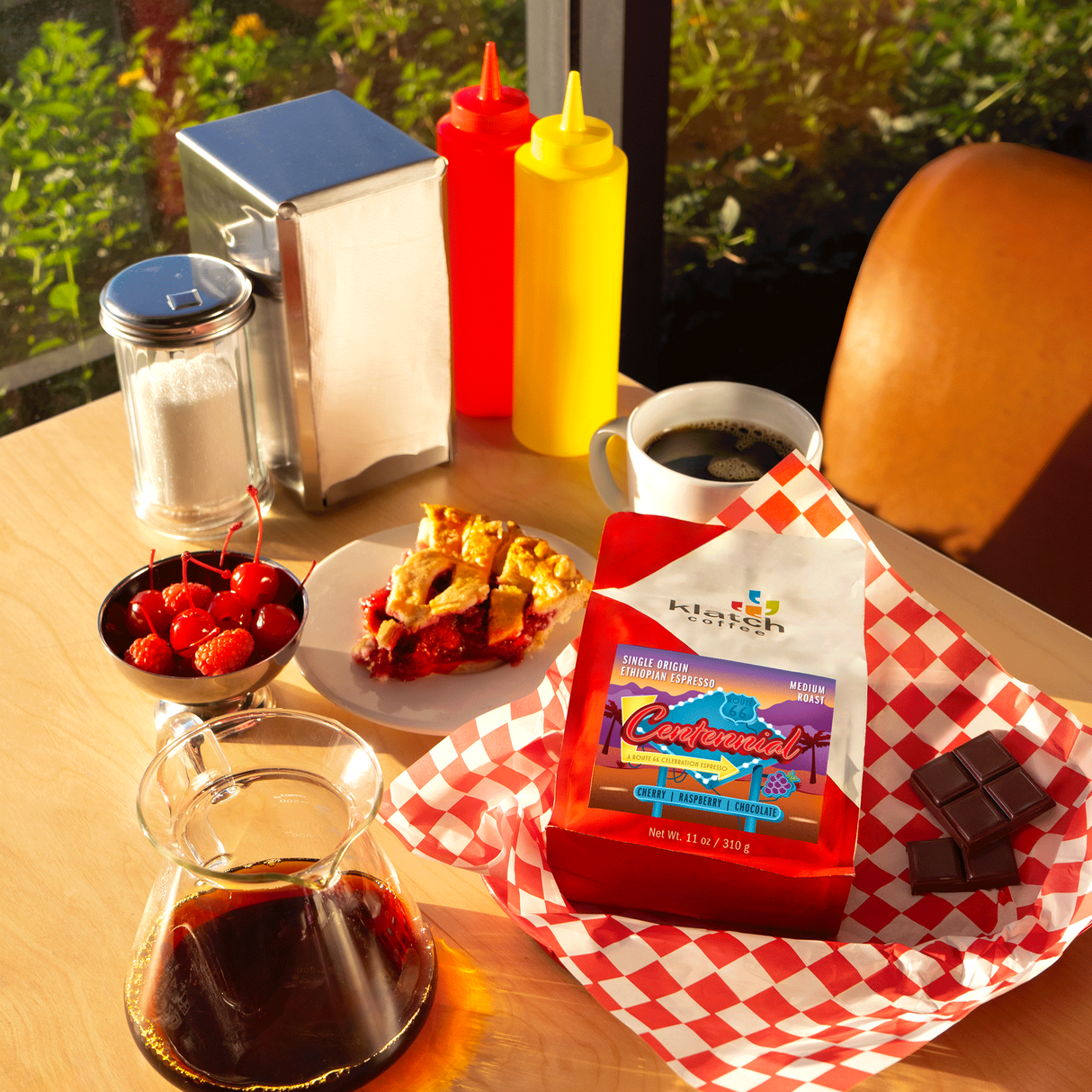 New Centennial Route 66 coffee bag on a diner table with a coffee mug, cherry pie, cherries/ raspberries in a bowl.