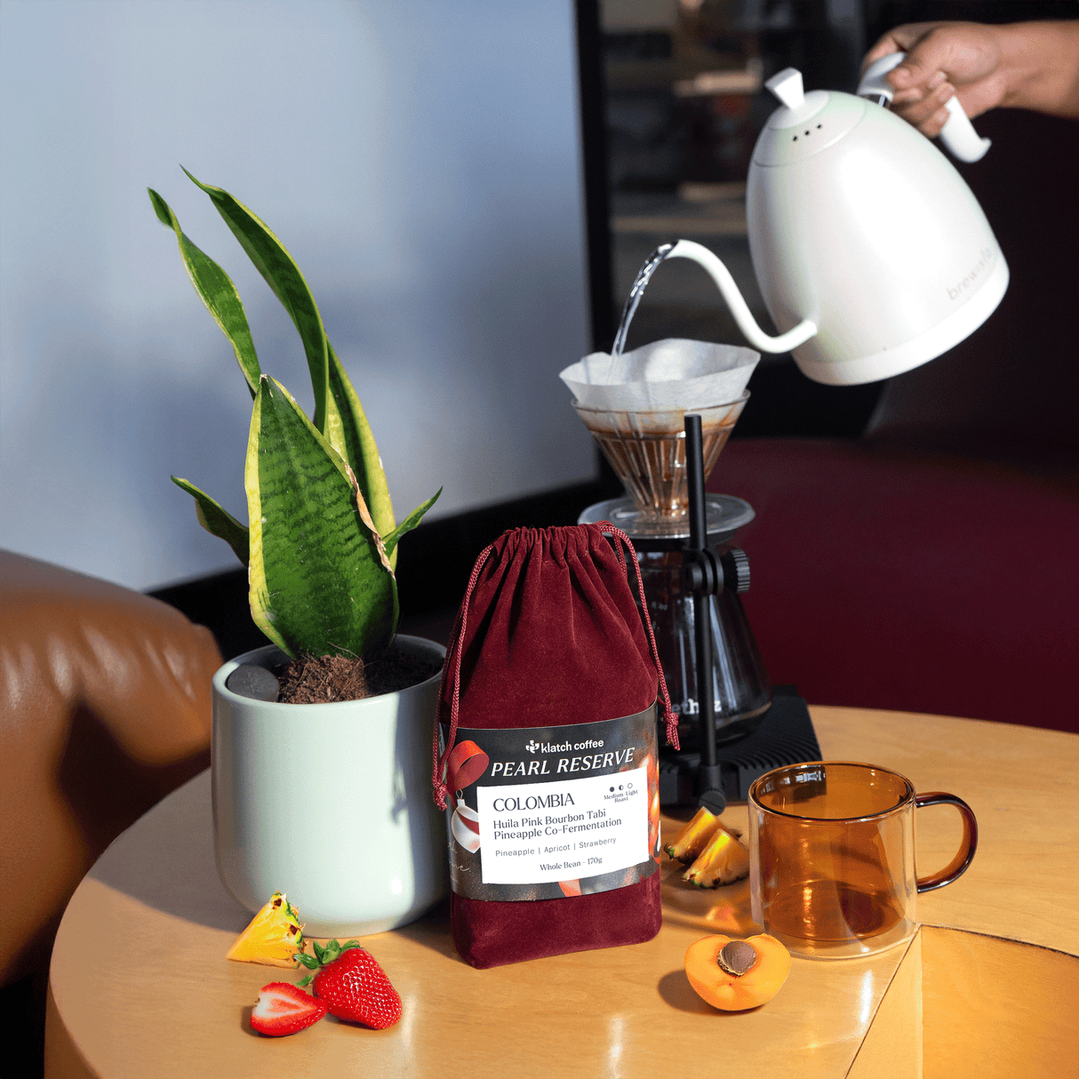 Red velvet drawstring coffee bag, with pour over, potted plant, with pour over, pine apple, strawberries and apricot and class amber coffee mug on wooden coffee table.