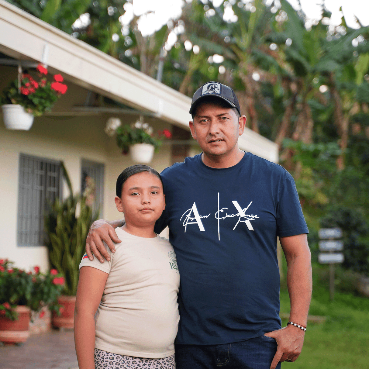 Two people standing together outdoors with a house and plants in the background