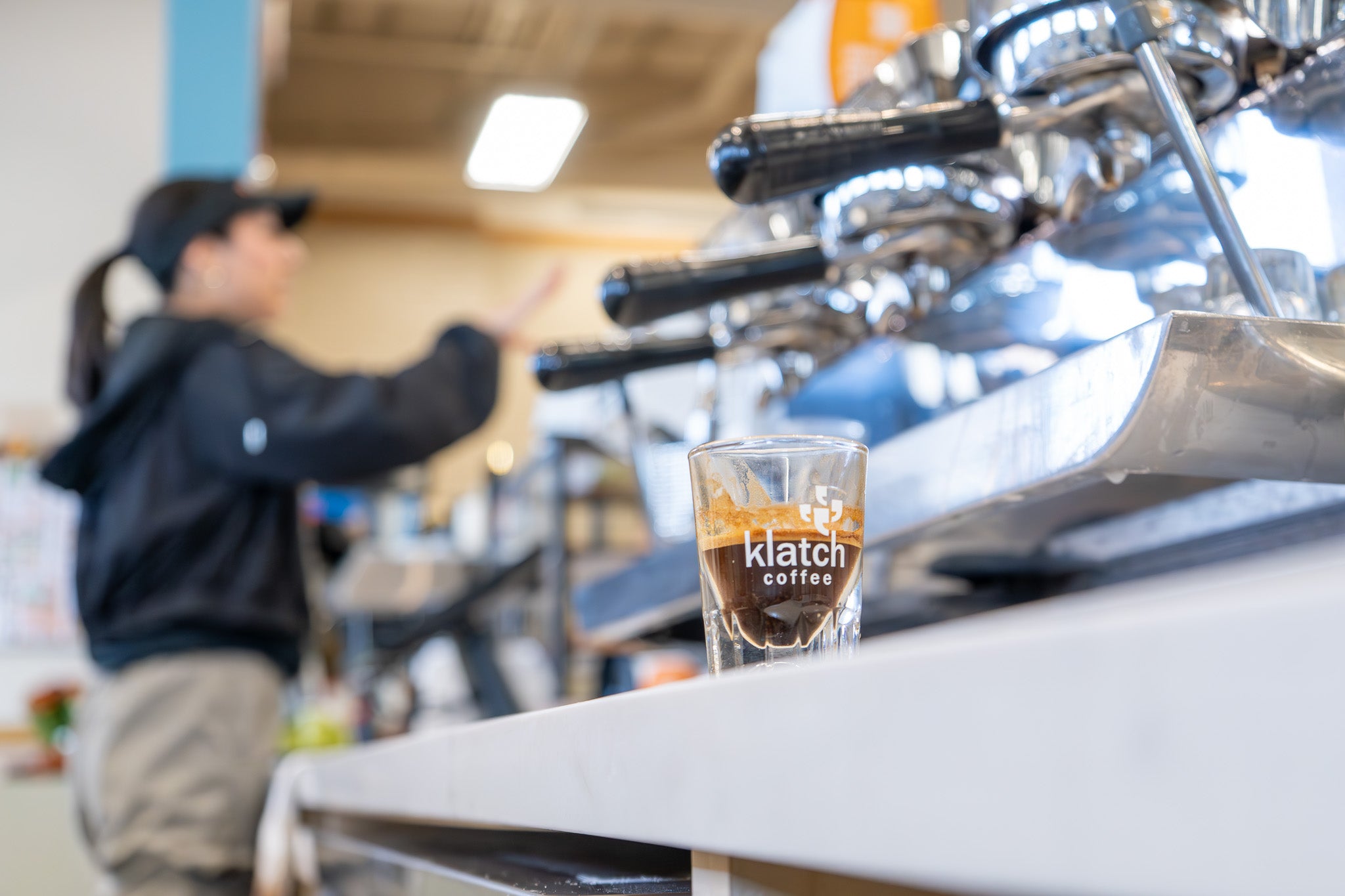 Shot of an espresso on the counter with the Klatch Coffee logo visible and a blurred barista in the background.