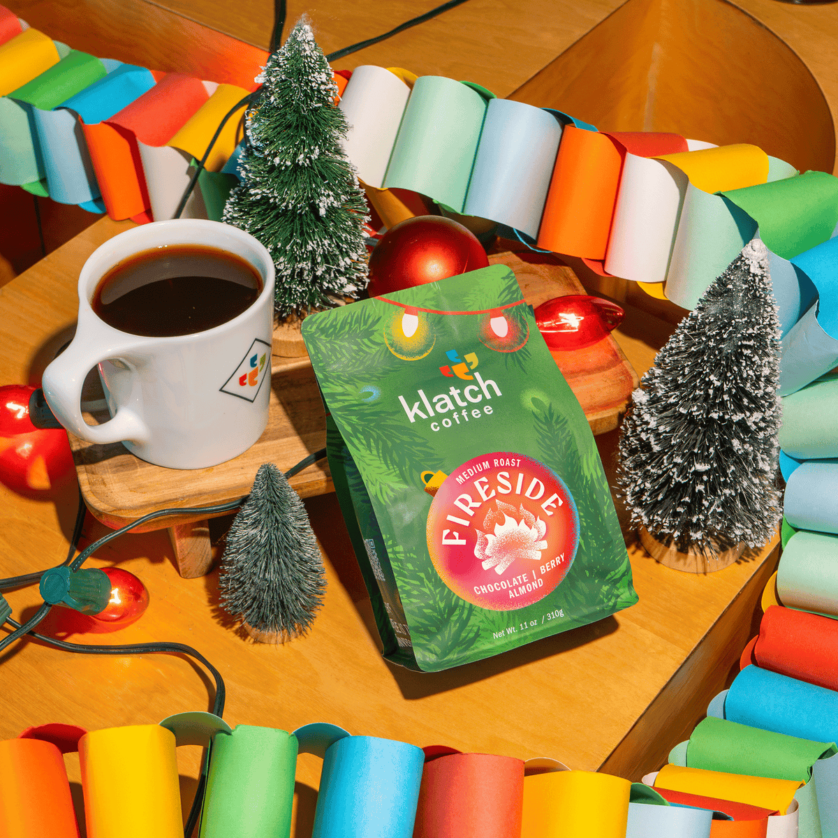 Klatch Coffee Fireside holiday coffee bag displayed beside a mug of coffee, miniature Christmas trees, colorful garlands, and festive lights.