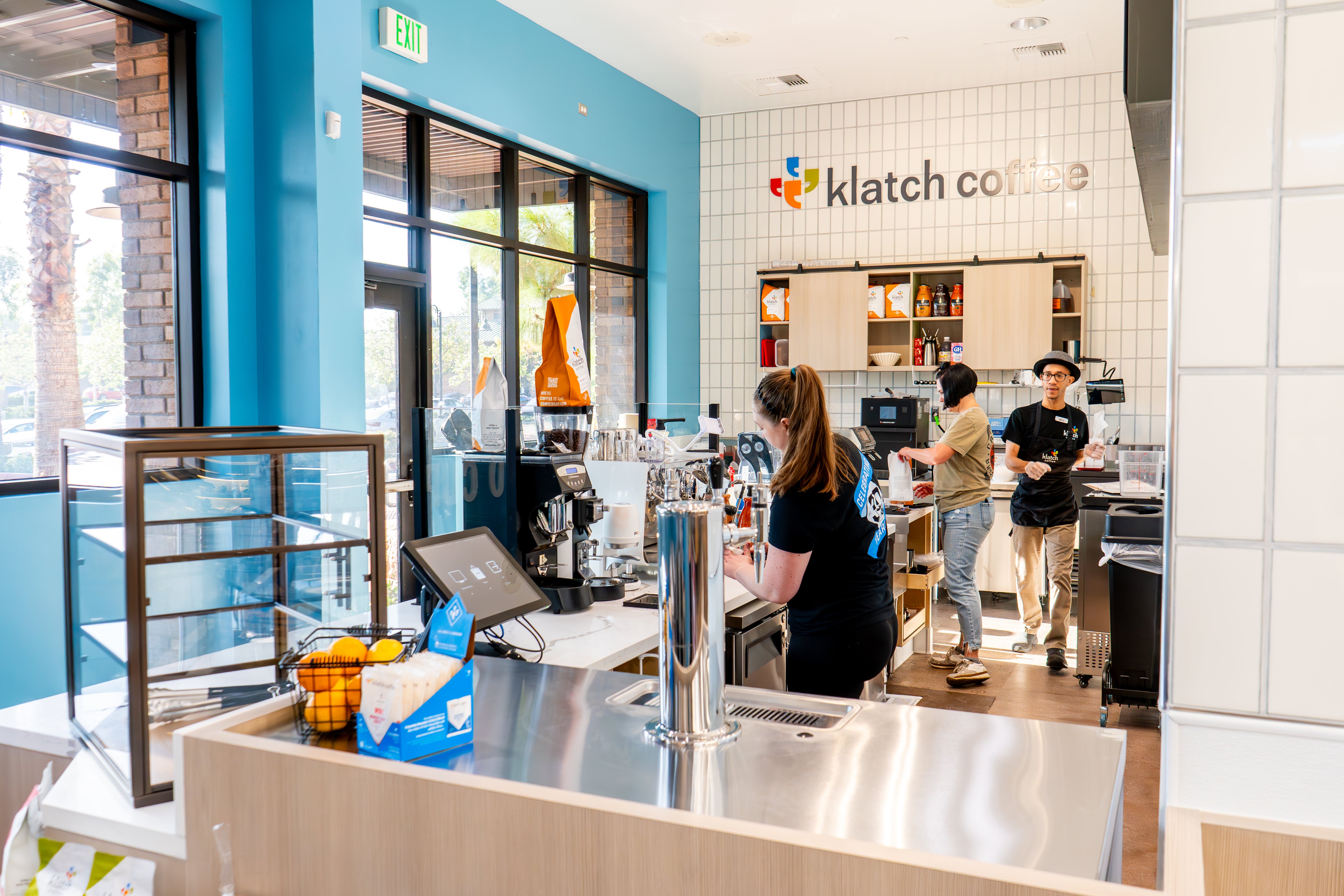 Three baristas preparing coffee behind the counter in a café, with the Klatch Coffee logo displayed on the wall.