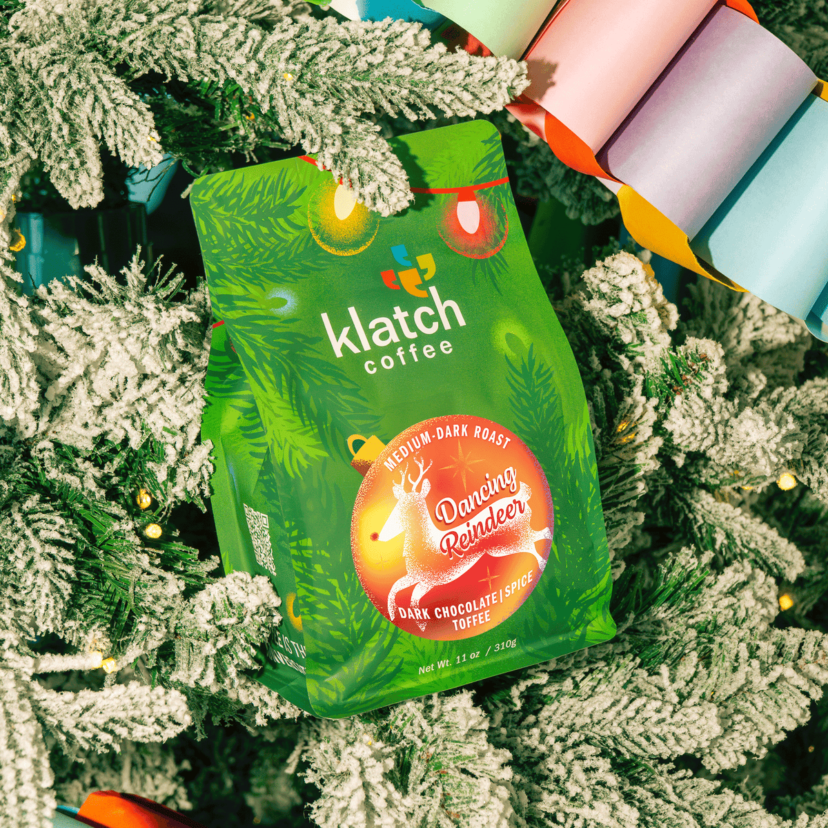 Klatch Coffee Dancing Reindeer holiday coffee bag featuring festive green packaging with a reindeer illustration, nestled among snow-covered Christmas tree branches, twinkling lights, and colorful paper garlands.