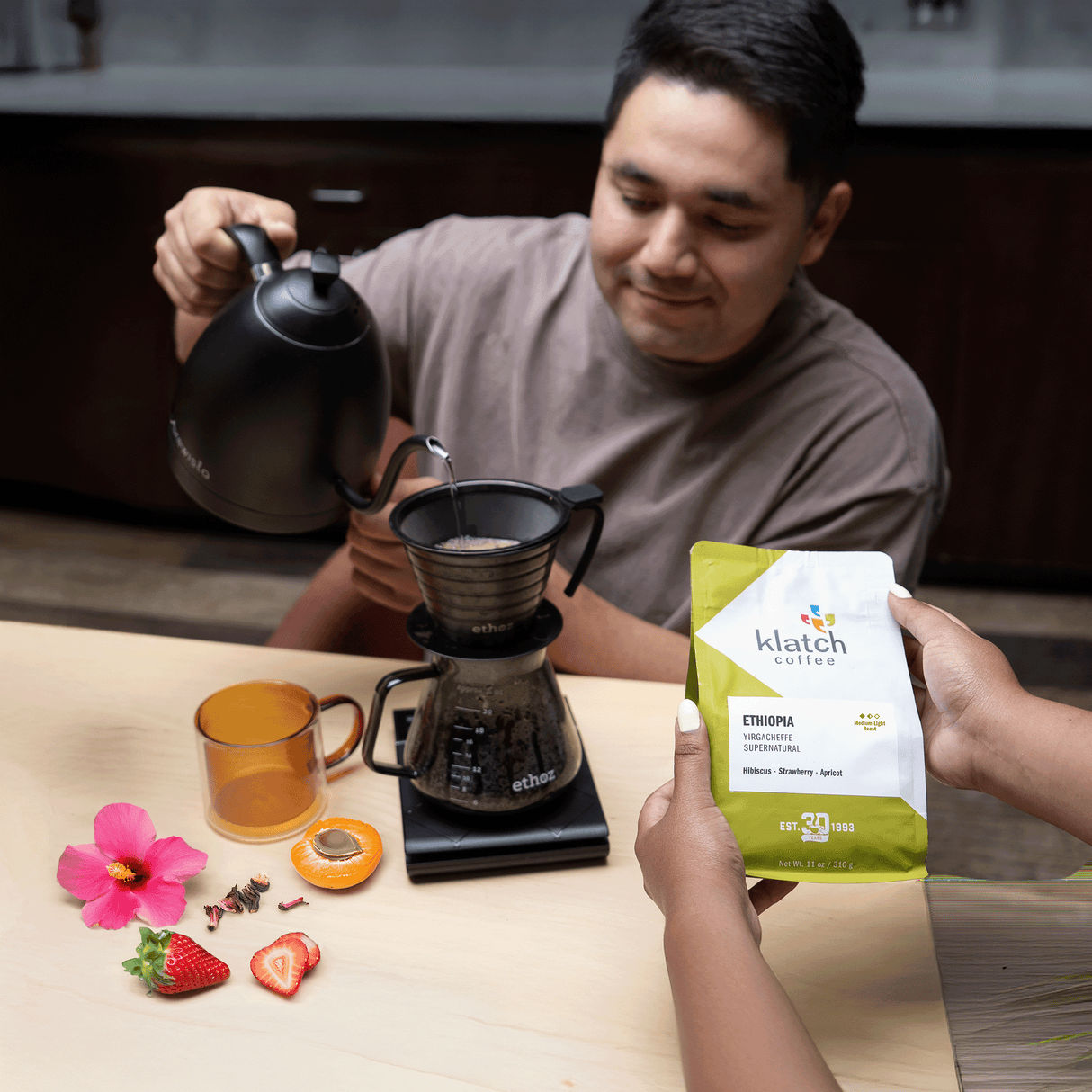 Man pouring hot water brewing pour over of with woman's hands holding bag of Ethiopia Supernatural.