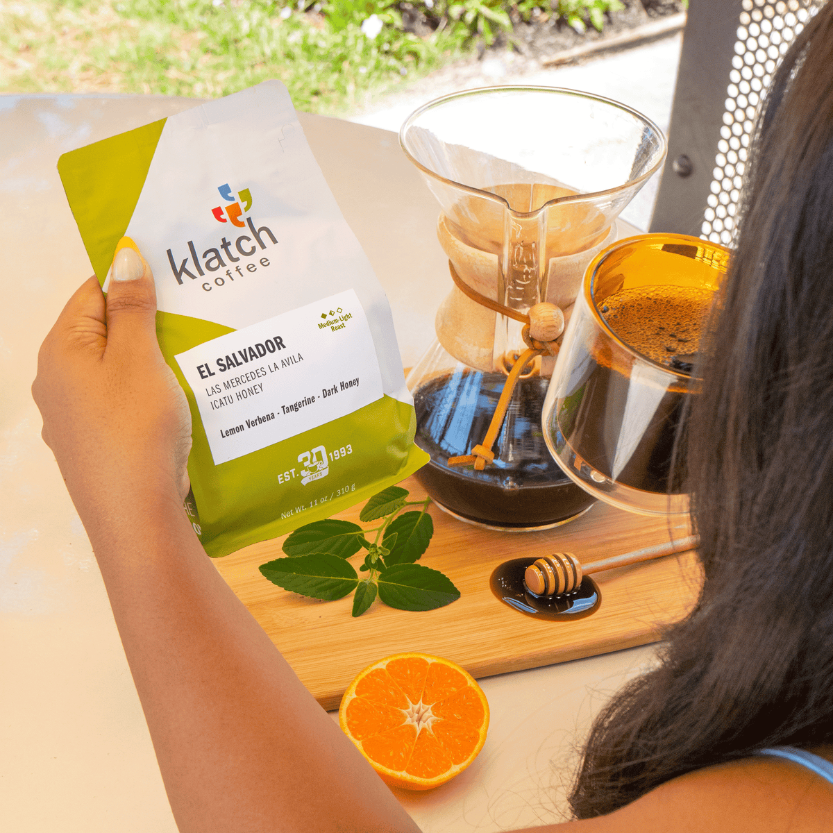 A person holds a bag of Klatch Coffee El Salvador coffee roast in a bright, sunny outdoor setting. A glass Chemex coffee maker and a glass cup of brewed coffee sit on a wooden board next to a honey dipper, a fresh orange slice, and mint leaves.