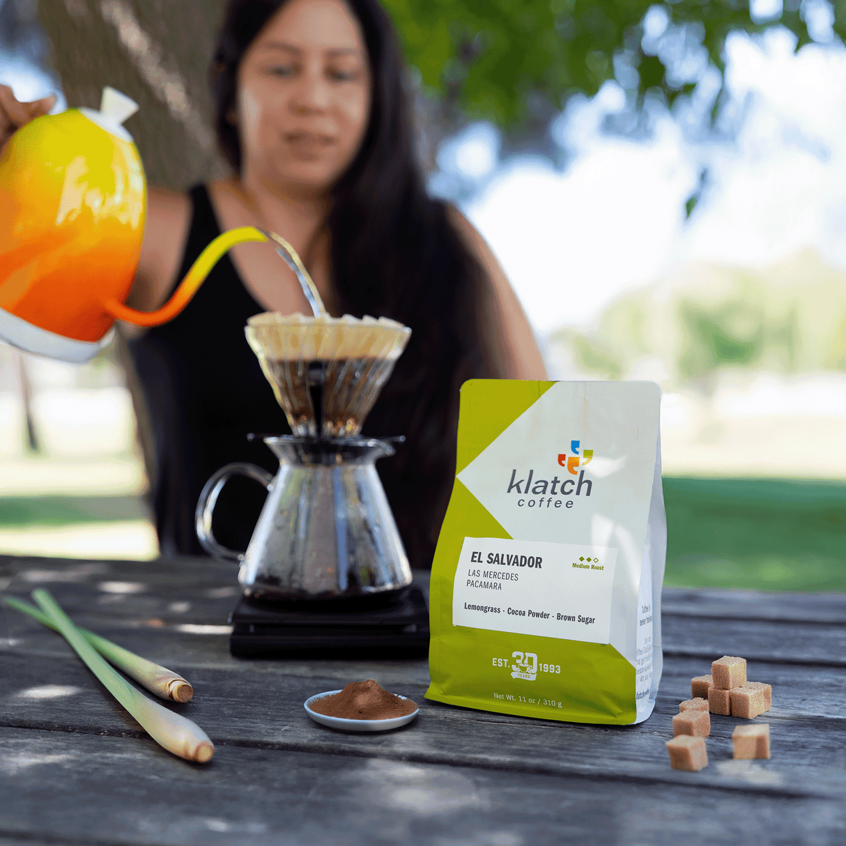 Women pouring hot water into brewing device with El Salvador Pacamara bag , lemongrass, cocoa powder and brown sugar cubes on wooden  picnic table outside.