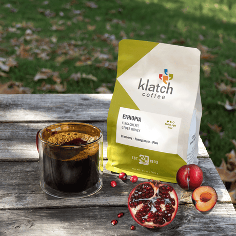 Klatch Coffee bag with a glass of coffee and fruits on a wooden surface outdoors.