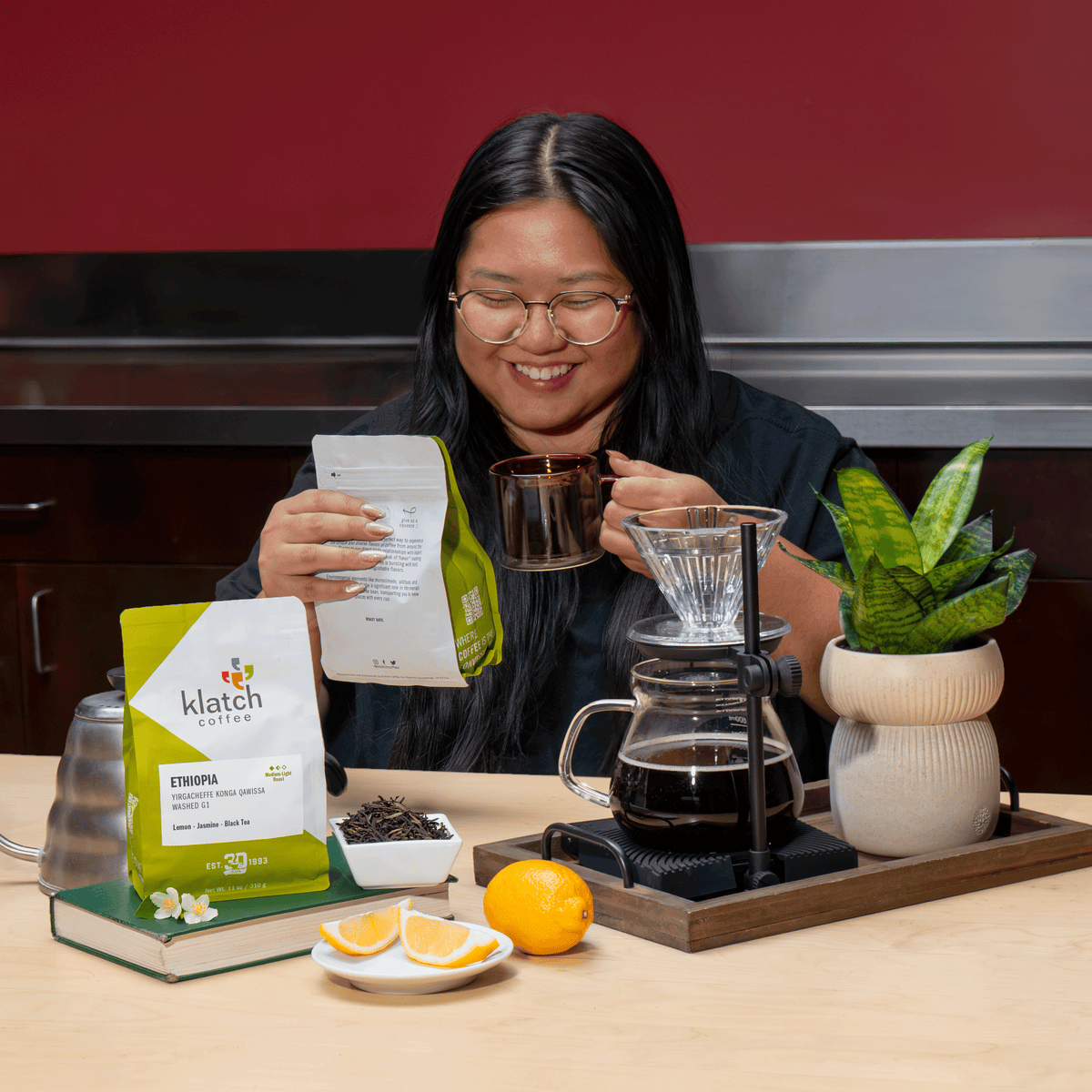 A woman with glasses is smiling while holding a bag of Klatch Coffee Ethiopia and a glass mug, standing behind a coffee bar setup with a pour-over dripper, glass carafe of coffee, oranges, and a potted plant.