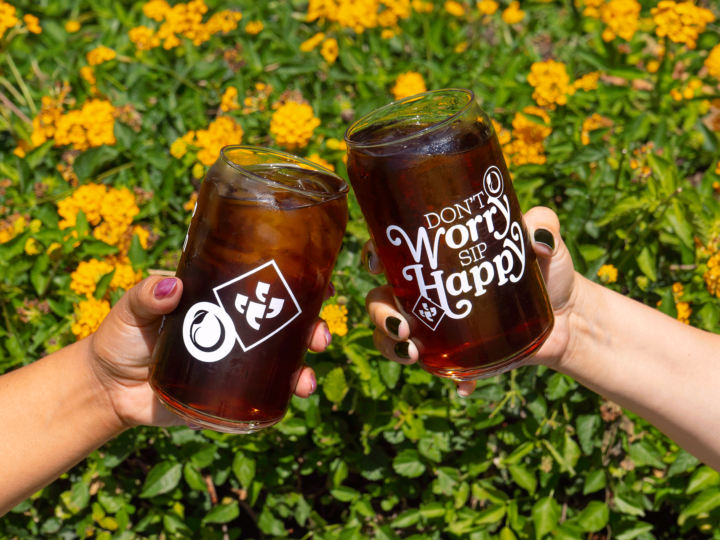 Close-up of two people clinking glass cups of cold brew in front of yellow flowers. One glass features the Klatch Coffee and Sprouts logos, while the other reads, "Don't Worry, Sip Happy."