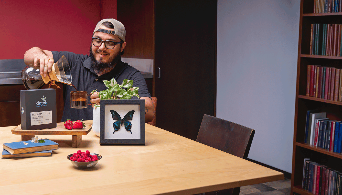 Man pouring coffee at a table with a framed butterfly, a plant, and a pearl coffee. Nearby are strawberries, jasmine flowers, and a bowl of raspberries.