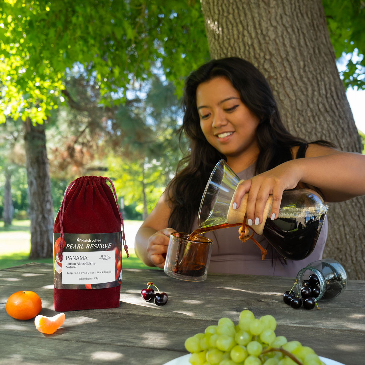 Woman pouring coffee into a glass at a picnic table with a bag of coffee and fruits.