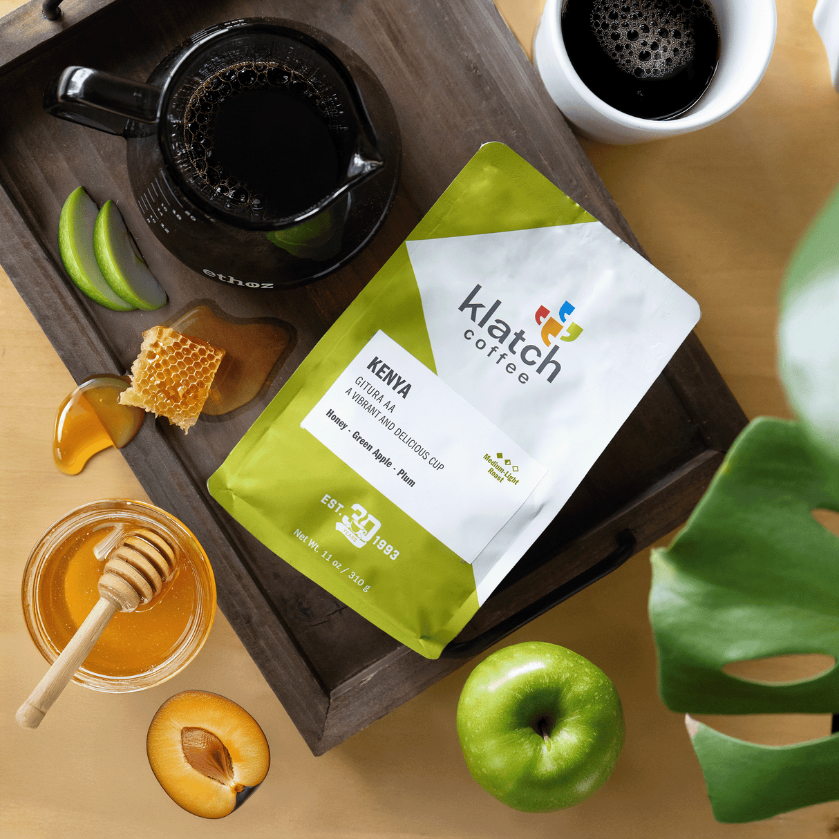 Klatch coffee Kenya Gitra AA bag on a wooden tray with green apple, honey, and plum set on a wooden table with a craft and mug of coffee.