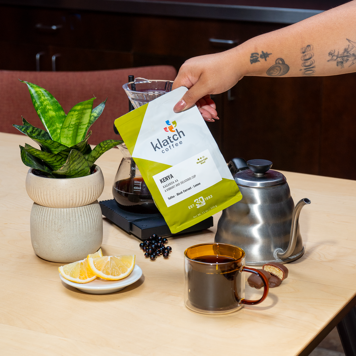 A hand holds a bag of Klatch Coffee Kenya Kaganda AA in front of a coffee setup on a light wood table, including a gooseneck kettle, brewed coffee in a glass mug, lemon slices, and a potted plant.