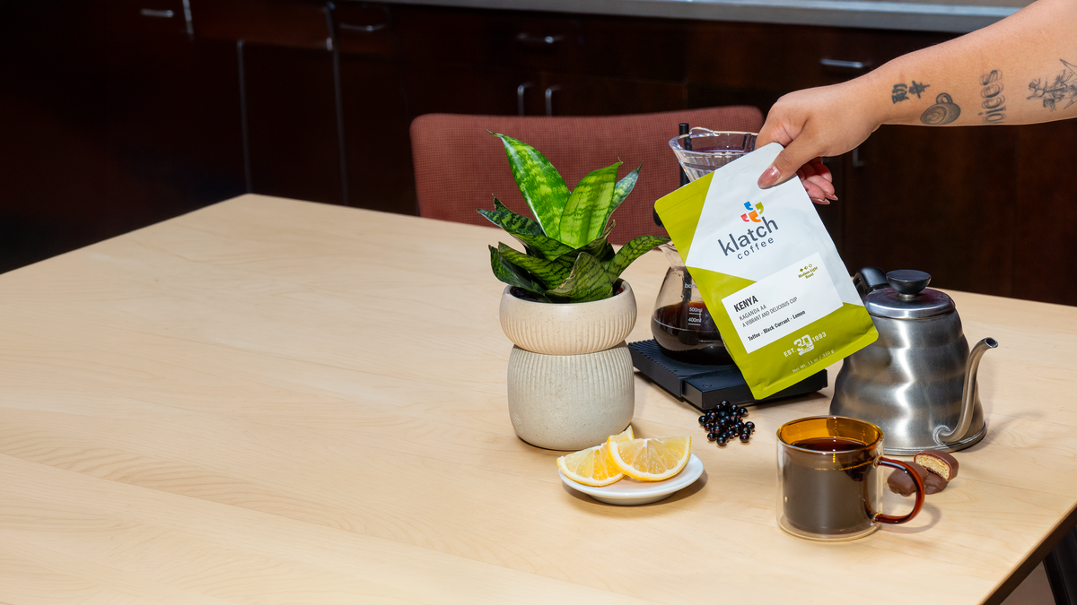A hand holds a bag of Klatch Coffee Kenya over a coffee setup on a wooden table, with a stainless steel kettle, glass mug of coffee, sliced oranges, and a potted plant in the background.