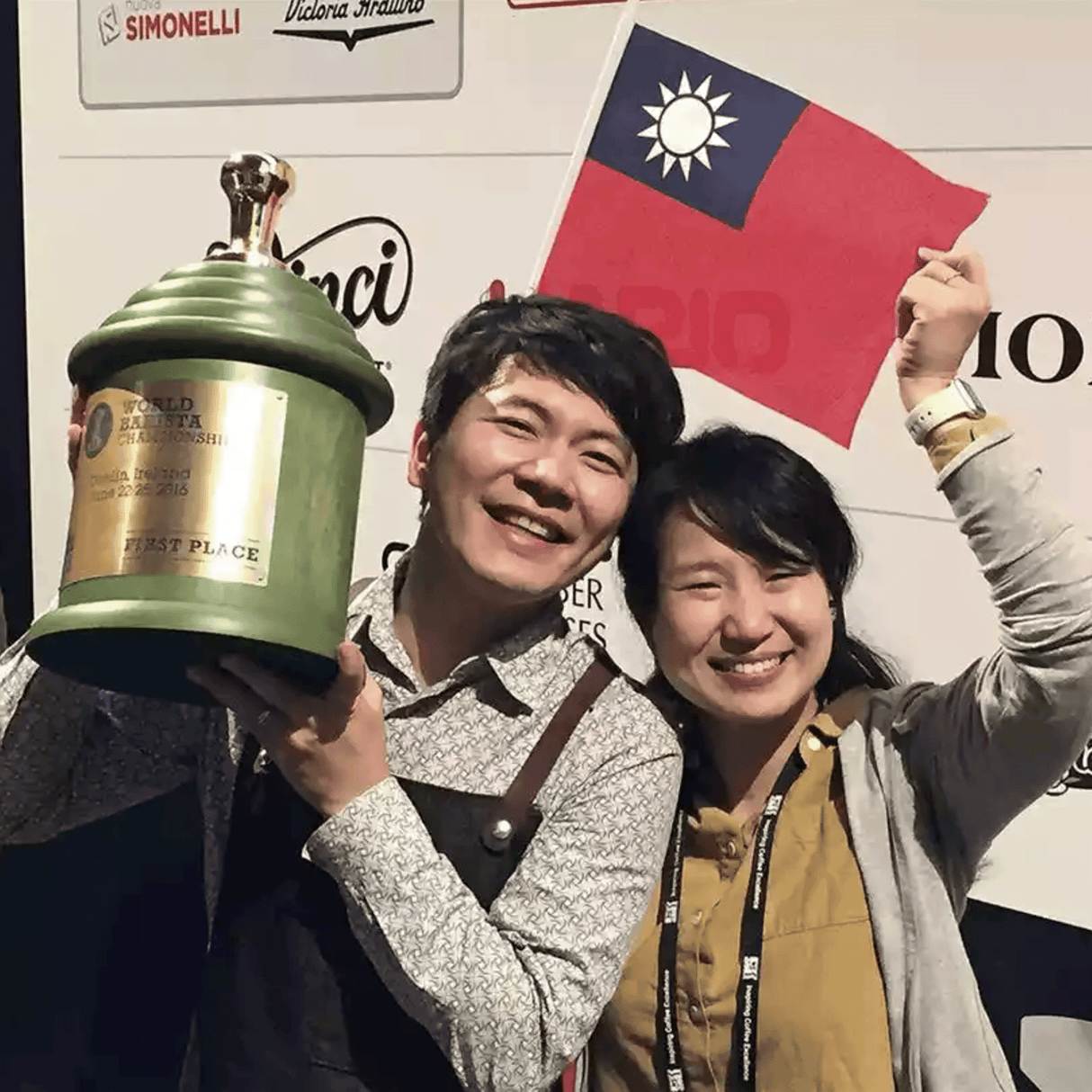 Photo of Berg Wu smiling and holding First place World Barista cylindar trophy with golden tamper on top next to a woman holding flag.