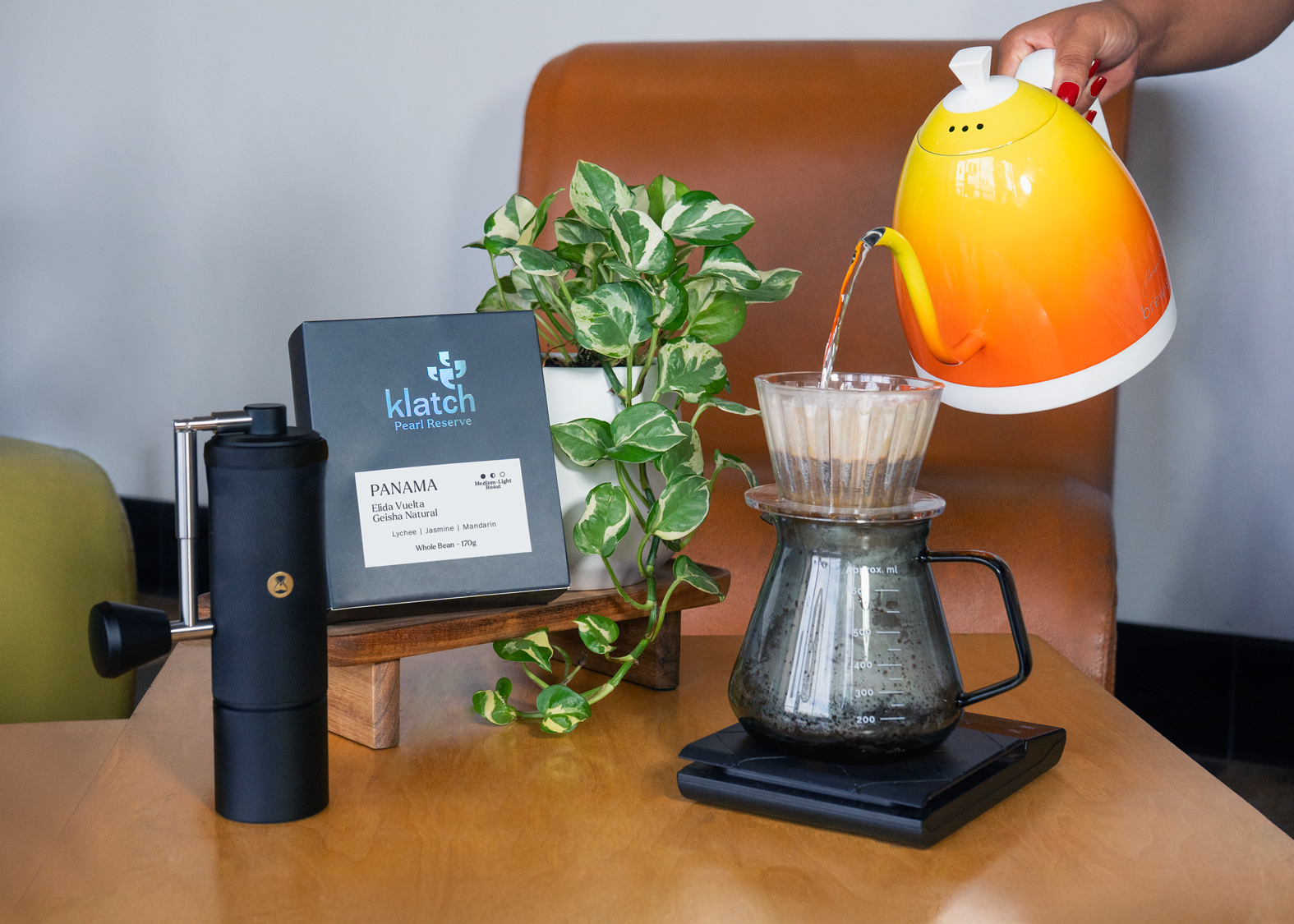 A person pours water from an orange kettle into a dripper over a coffee server, with a black grinder, a bag of Panama Elida Vuelta Pearl Reserve, and a green plant arranged on a table in front of a brown couch.
