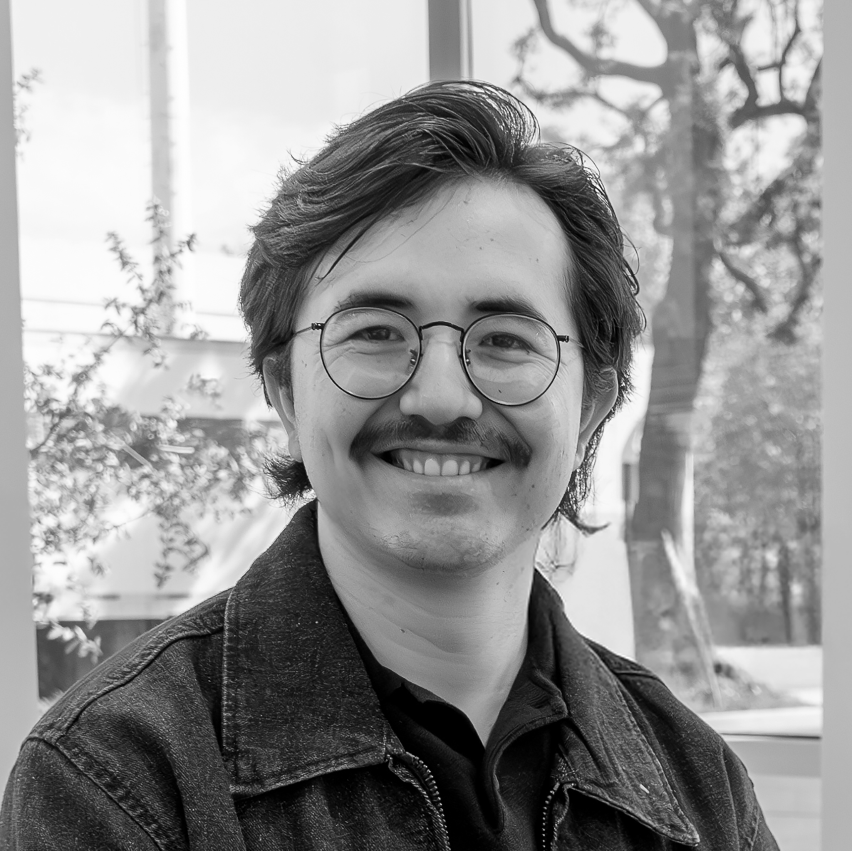 Black and white photo of a person with glasses and a jacket, standing indoors with windows in the background.