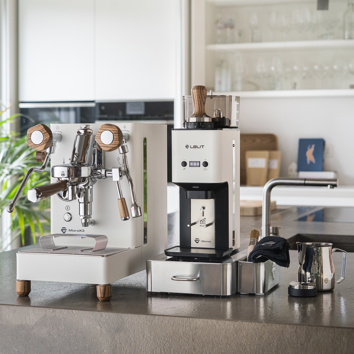A white LELIT MaraX3 Espresso Machine set with a matching William Coffee Grinder and accessories, arranged on a kitchen countertop in a bright, modern kitchen.