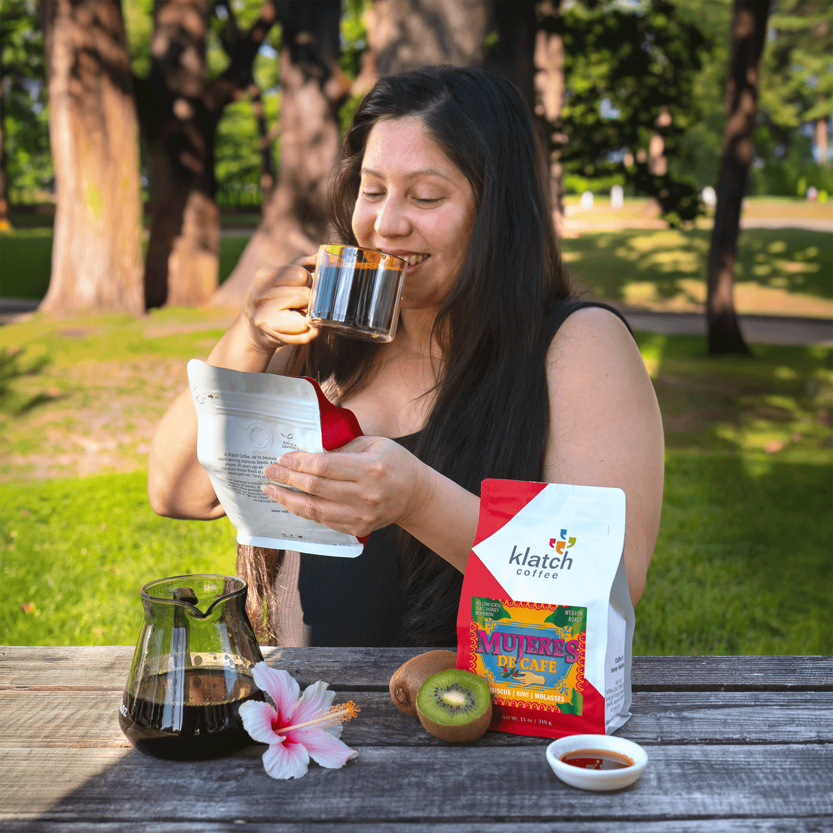 Woman enjoying a cup of coffee outdoors with a Klatch coffee Mujeres De Cafe bag and other items on a table.