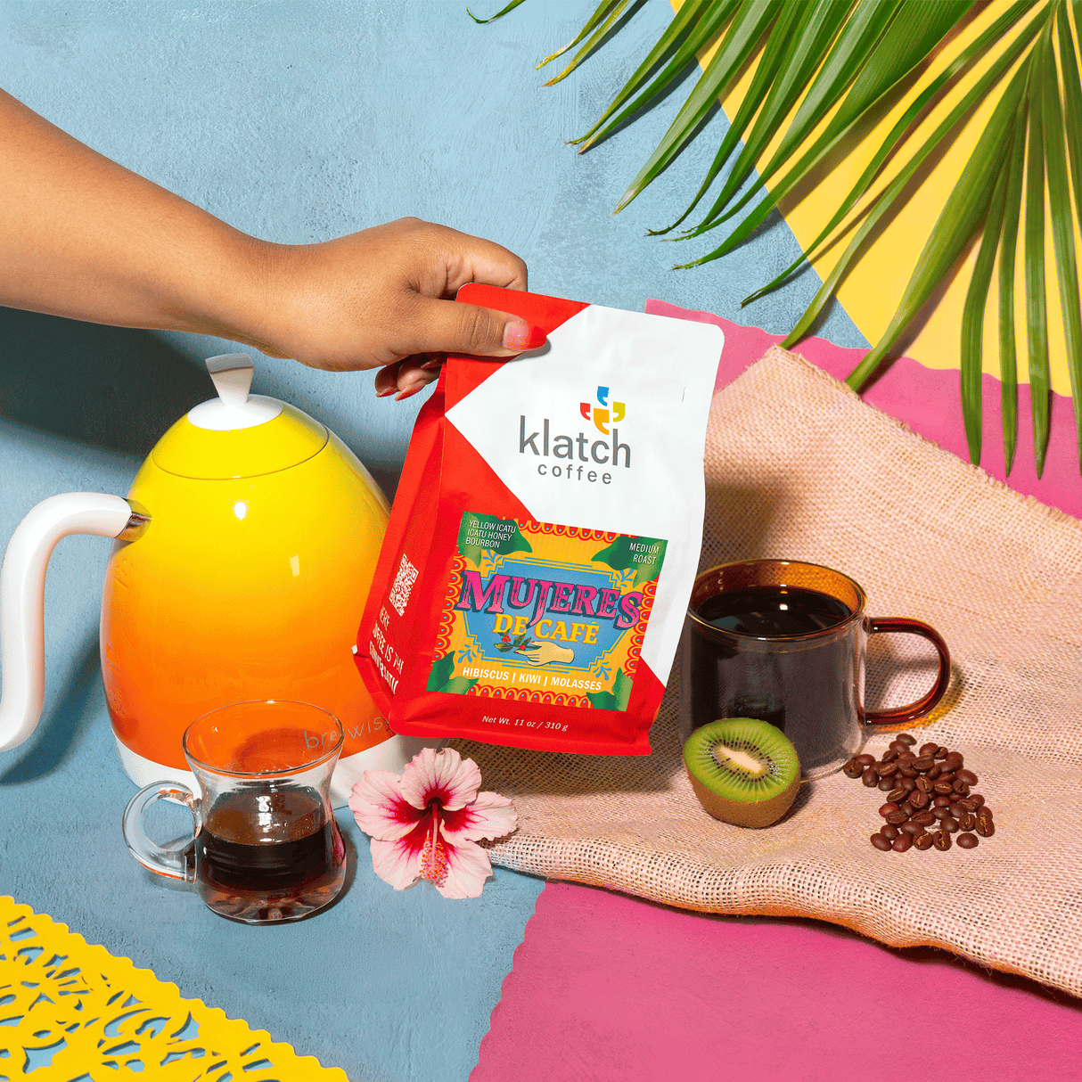 Hand holding a Klatch coffee bag with a colorful background featuring a kettle, coffee cups, and coffee beans.