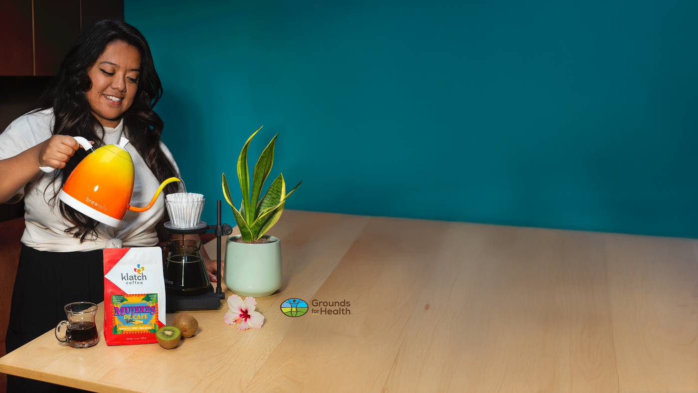 Woman pouring Mujeres De Cafe coffee into a cup on a wooden table with a teal wall background.