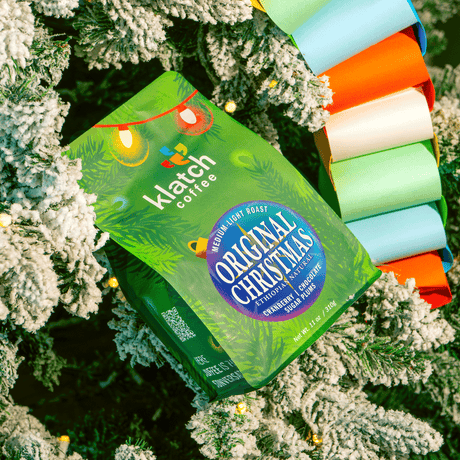 Klatch Coffee Original Christmas holiday coffee bag featuring festive green packaging with colorful Christmas lights, nestled among snow-covered Christmas tree branches, twinkling lights, and a vibrant multicolored paper garland.
