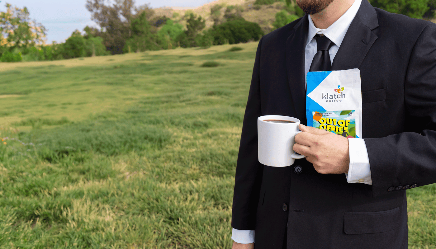 Man in a suit holding a Klatch coffee Out of Office bag of coffee and a mug outdoors.