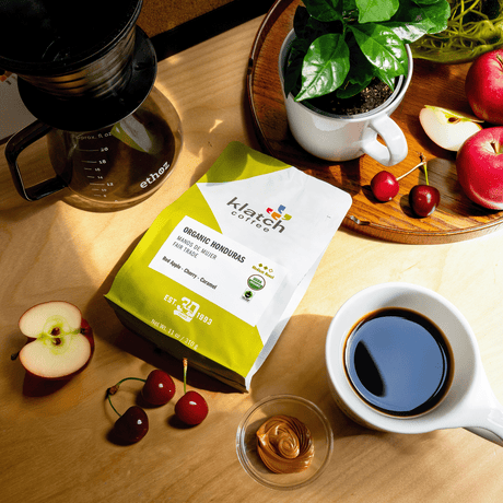 Organic Honduras Mano De Mujer Fair Trade coffee bag on wooden table, glass ethoz pour-over with white mug with coffee. Fruit of flavor notes of Coffee plant and  red apples, one cut in wedge with two cherries on tray with half apple and cherries laying on table at the bottom of the bag next to glass bowl of caramel and mug of coffee.