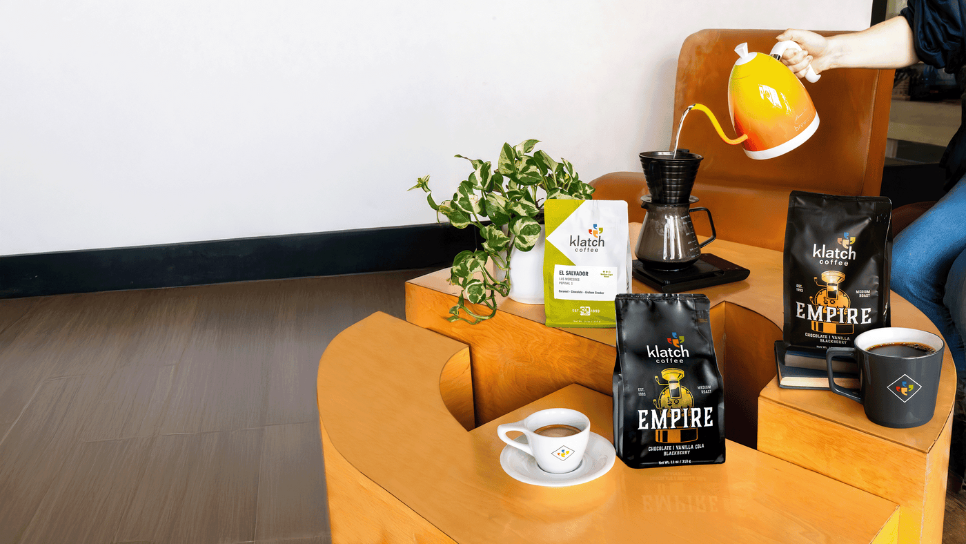 Person pouring hot water into brewing device on coffee table with three coffee bags, coffee mug and espresso.