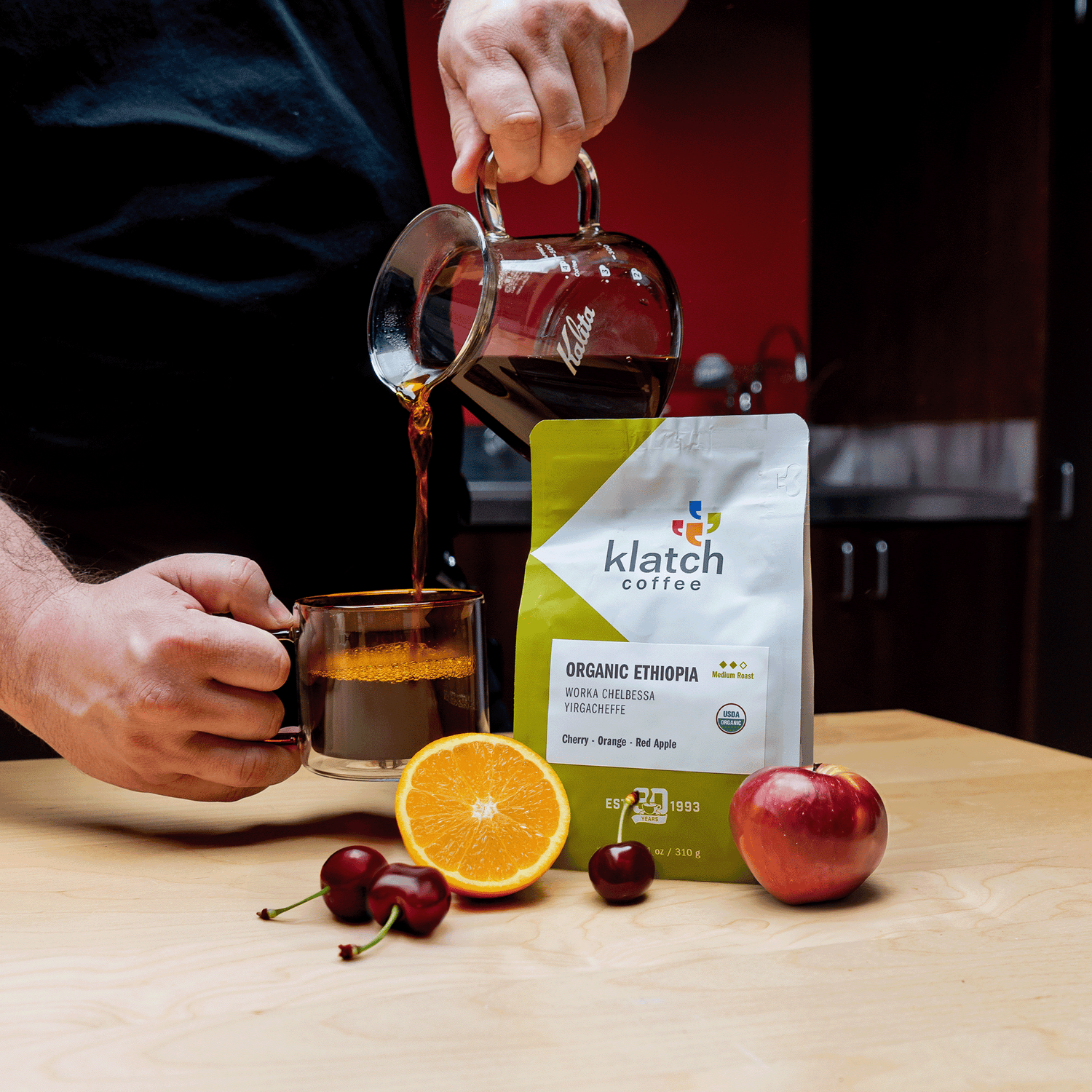 Organic Ethiopia coffee being poured by man into glass amber mug over wooden table, next to bag of coffee with fruit, half orange, cherries and red apple.