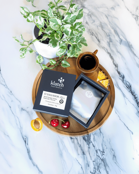 Klatch coffee El Salvador Jasal Orange Geisha - 94pts Coffee review badge package on a wooden tray with a cup of coffee, plant, and fruit on a marble background