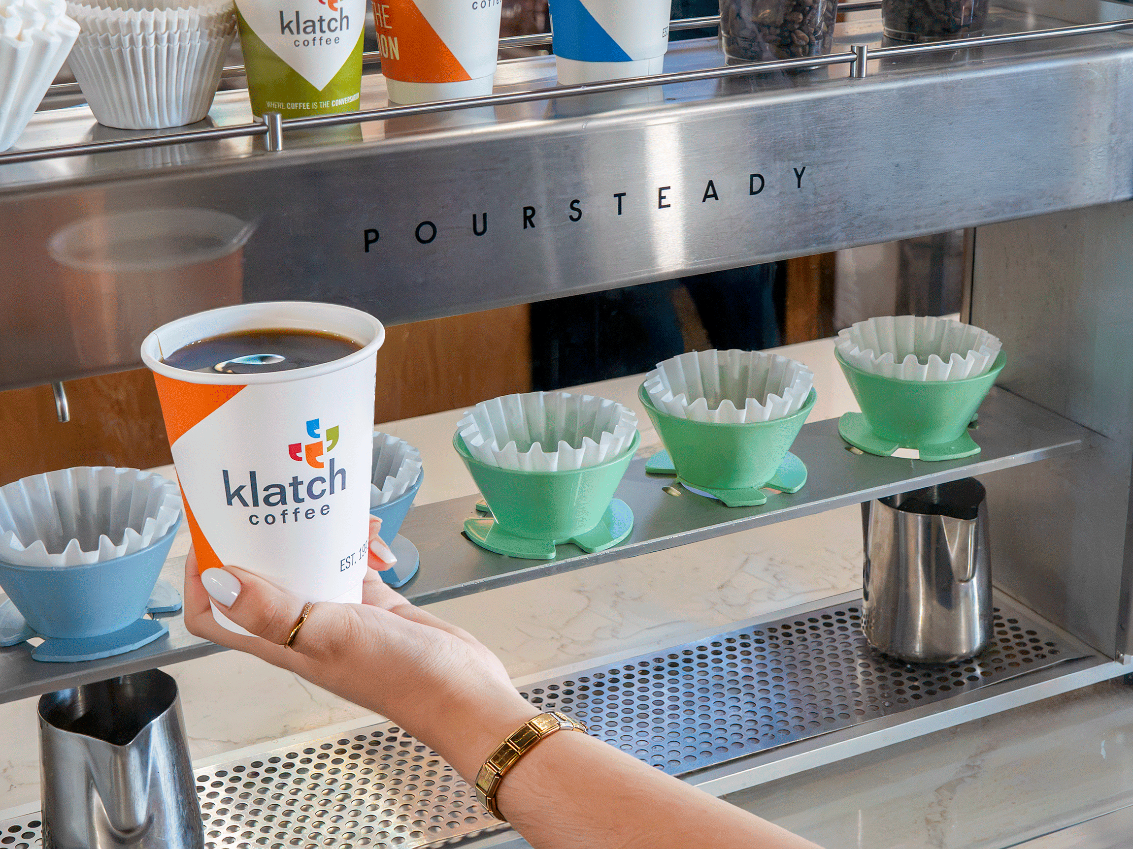 Hand holding an orange Klatch Coffee cup in front of a Poursteady automated pour over brewer.