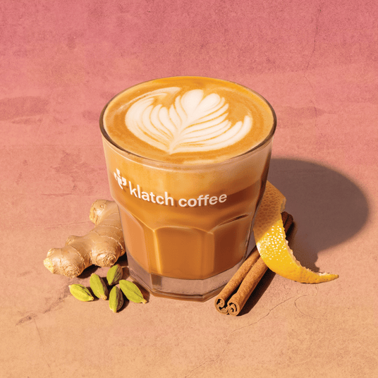 A latte in a clear glass cup with the Klatch Coffee logo, surrounded by fresh ginger, cinnamon sticks, green cardamom pods, and a curled lemon peel.