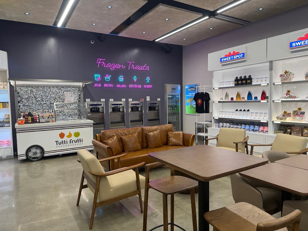 Interior of Tutti Frutti featuring frozen yogurt machines, a toppings counter, merchandise displays, and a modern seating area with tables, chairs, and a leather sofa.