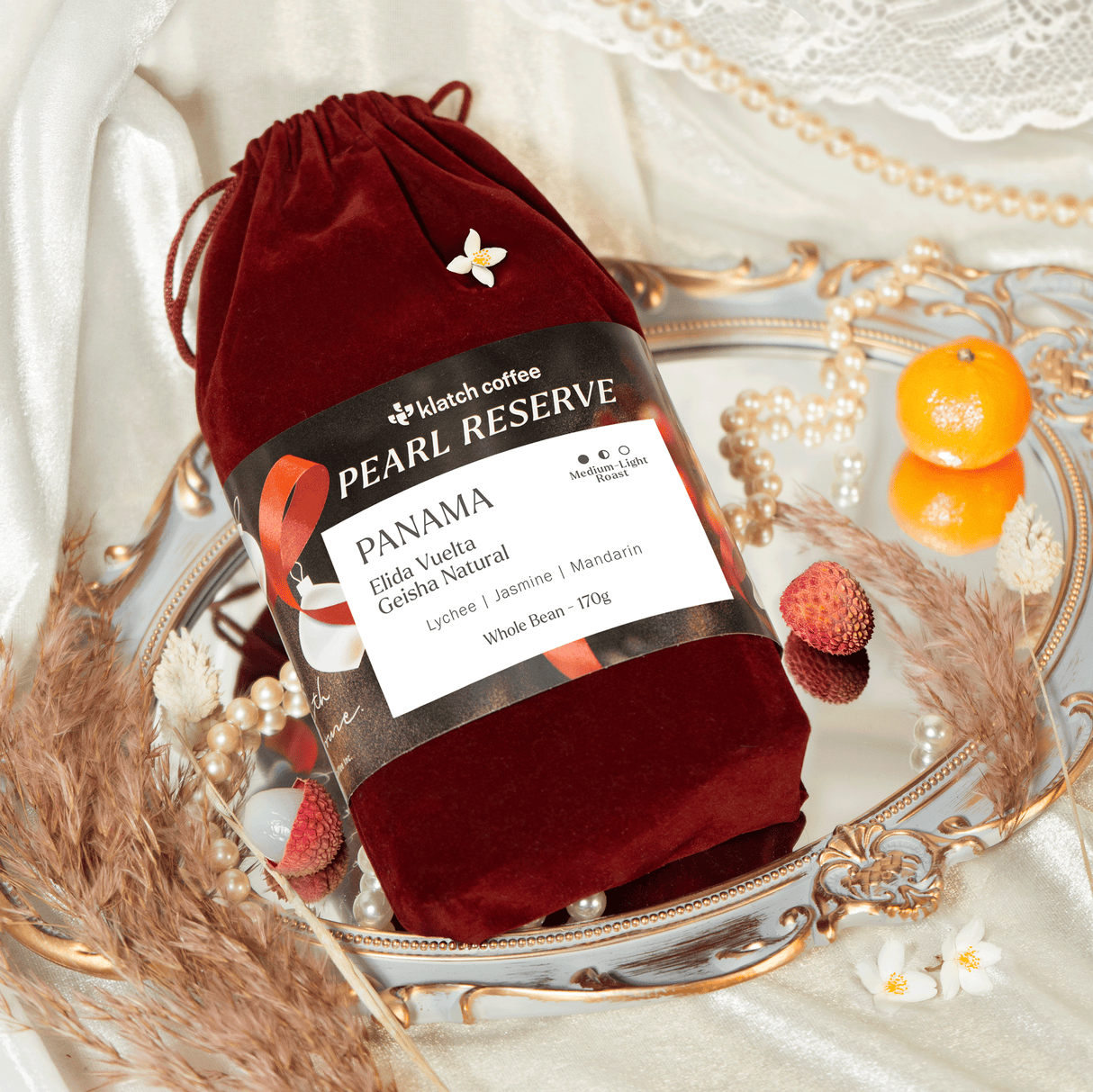 Red velvet drawstring bag with Panama Elida Vuelta Geisha coffee label on a decorative mirror tray with fruits.