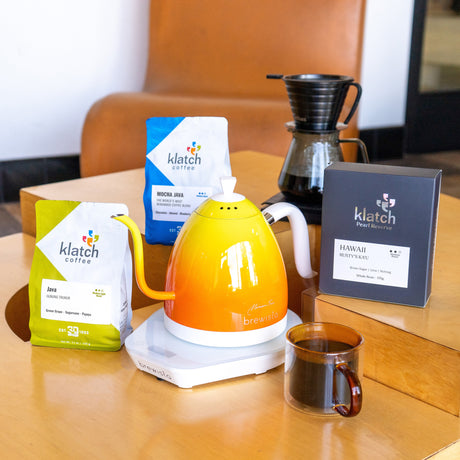 Asian Klatch Coffees of Mocha Java, Java and Hawaii Ka'u with Brewista kettle on top of wooden coffee table with glass pour-over and glass mug with coffee.
