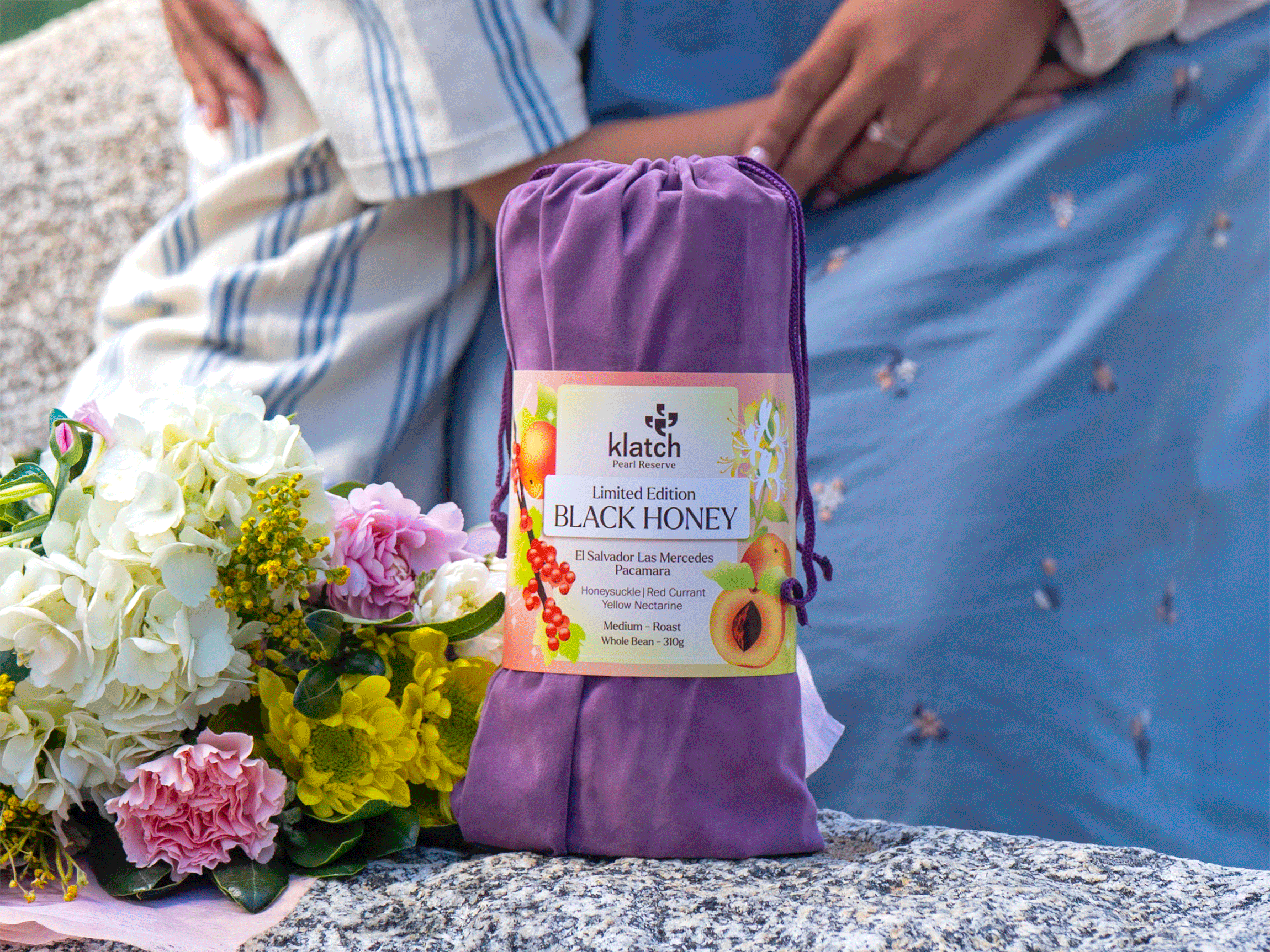 Purple velvet drawstring bag with a wraparound label reading “Limited Edition Black Honey El Salvador Las Mercedes Pacamara,” placed on a rock beside a bouquet of flowers.