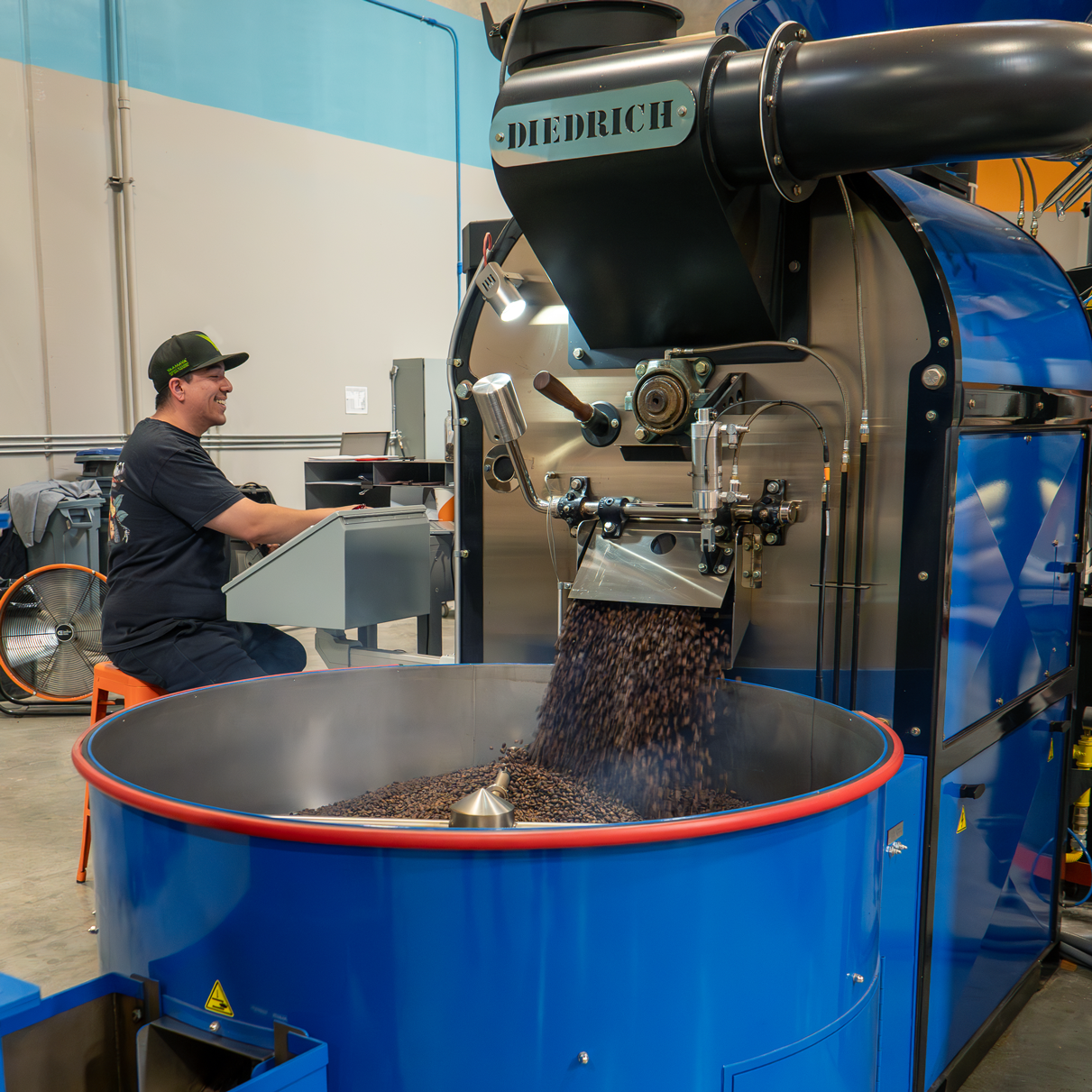 Person operating a coffee roasting machine with ' Diedrich' branding in a warehouse setting.