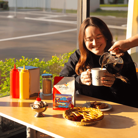 Diner setting with women pouring coffee into mug with another women holding mug sitting and a Centennial bag of coffee. With chocolate, cherry pir, ice cream scoop w/ cherry on top on a wooden table.