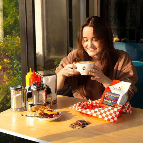 Woman enjoying a latte at a table with a a slice of cherry pie, and a bag of Centennial Espresso.