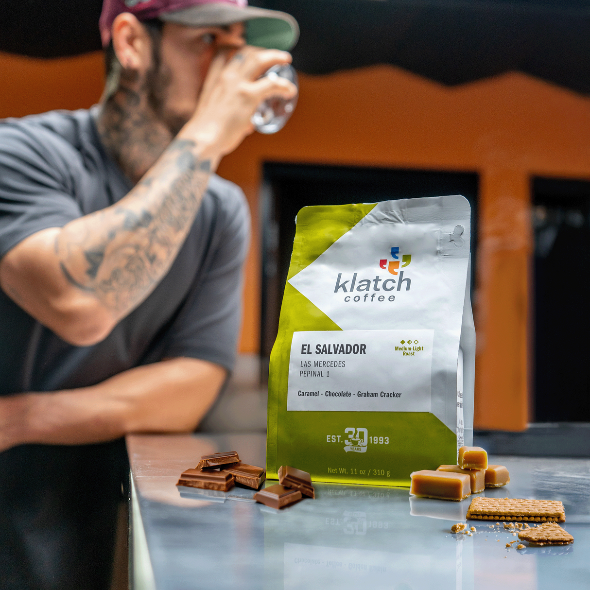 Man drinking from a glass with a Klatch Coffee El Salvador Las Mercedes Pepinal 1 bag with caramel candies, chocolate stacked and graham cracker on a table.